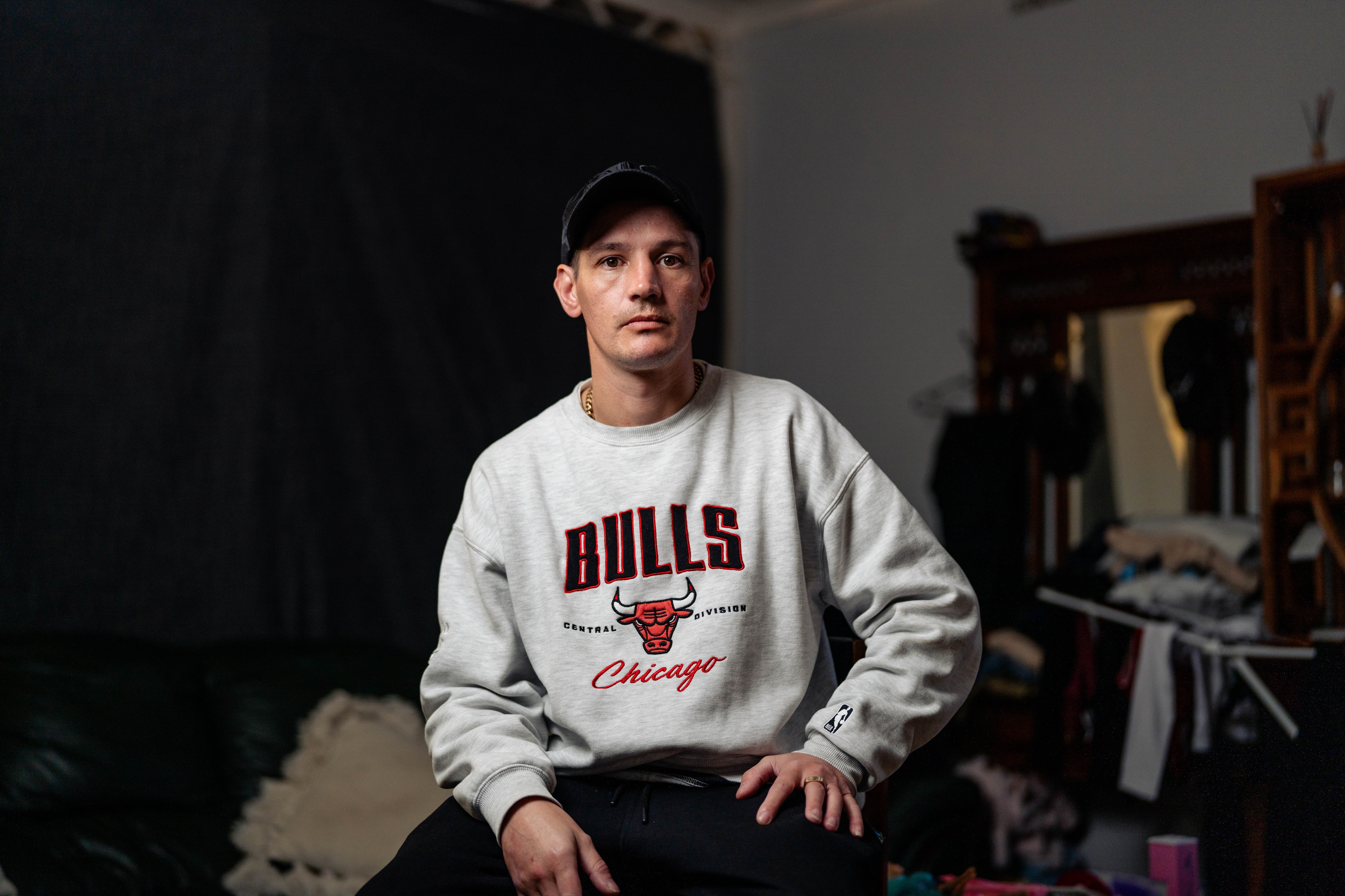 Shannon, wearing a grey jumper and a black hat, sits on a stool in a dimly lit living room with a serious expression.