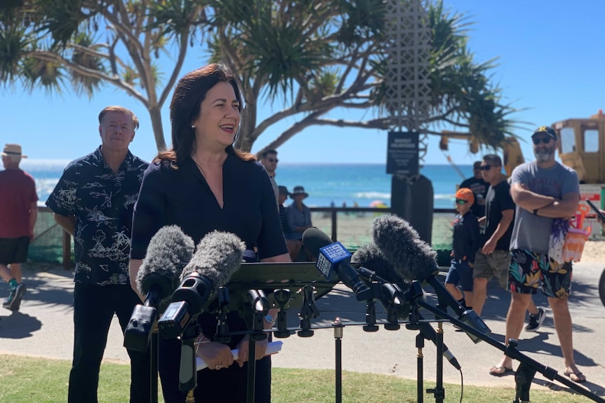 Annastacia Palaszczuk stands in front of microphones with Wayne 'Rabbit' Bartholomew behind her at the beach.