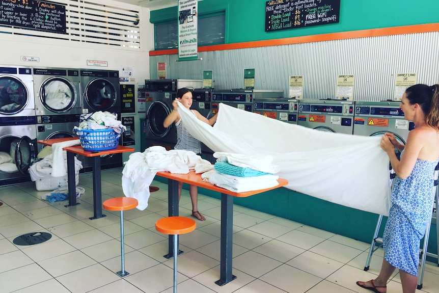 Two women in a laundromat working together to fold a white sheet.