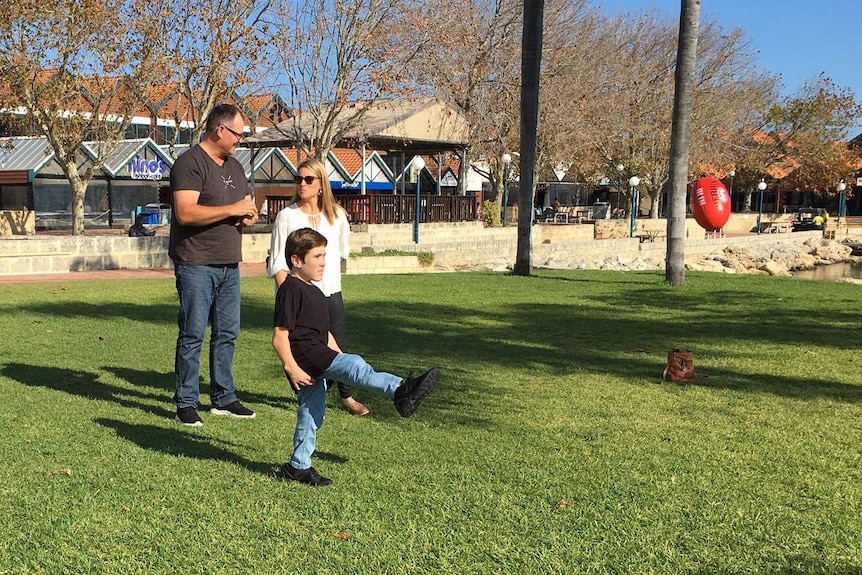Coby Antonio, who has Morquio A syndrome, kicks a football on a patch of grass with his parents standing behind him.