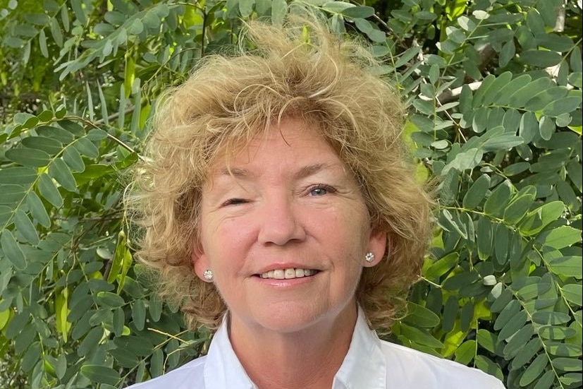 A woman with blonde hair stands in front of a tree and smiles