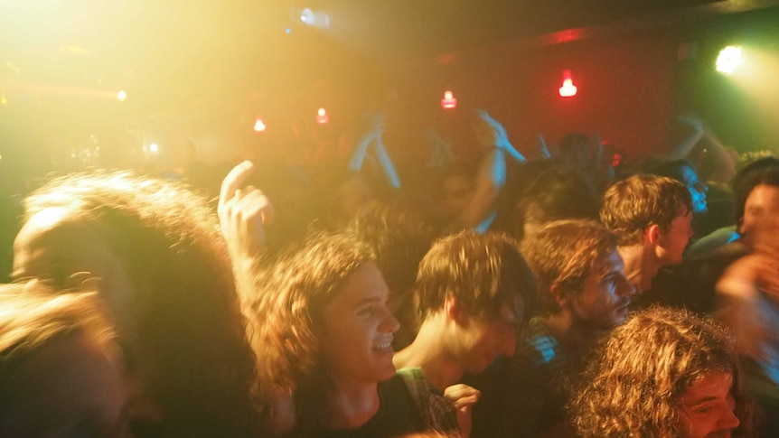 A picture of a bandroom with a crowd packed in tight, dancing under yellow and red and green lights.