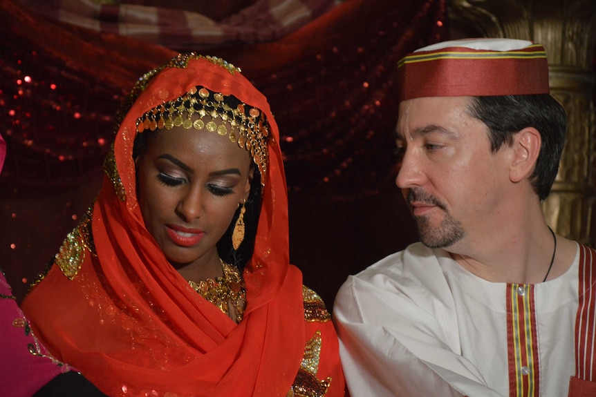 A couple wearing red and white traditional dress, with the man looking lovingly at the woman.