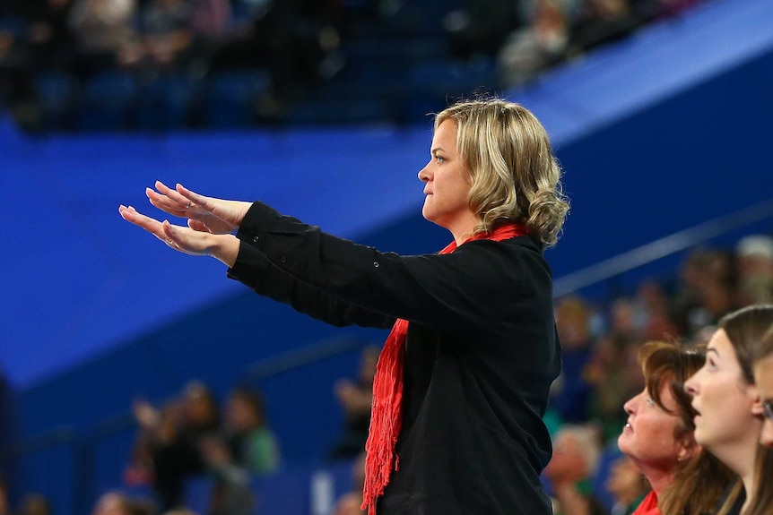 A netball coach stands facing the court, with her hands outstretched palms down to calm her team.