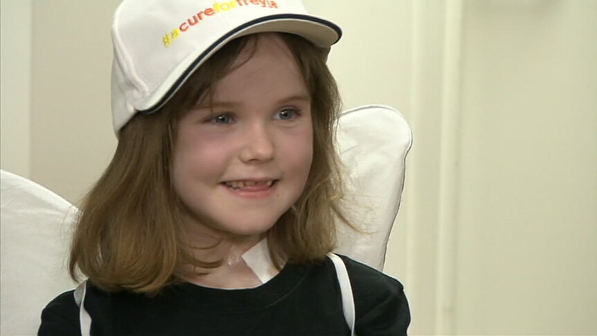 A girl wearing angel wings, and a cap with the words #acureforfreyja smiles while being interviewed after her surgery.