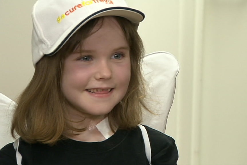 A girl wearing angel wings, and a cap with the words #acureforfreyja smiles while being interviewed after her surgery.