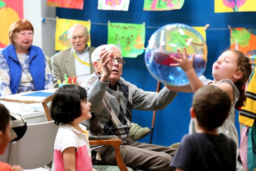 An elderly man and a young girl play with an inflatable ball in a classroom.