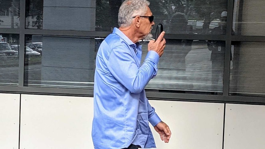 A greying man in a blue shirt walks by several windows attempting to cover his face.