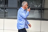A greying man in a blue shirt walks by several windows attempting to cover his face.