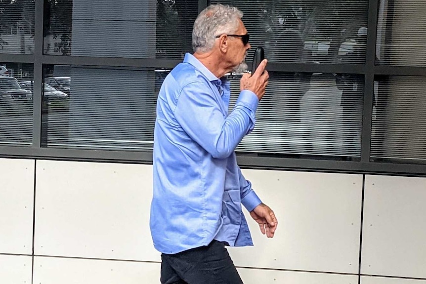 A greying man in a blue shirt walks by several windows attempting to cover his face.