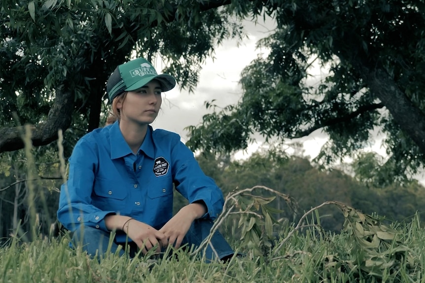 Cindy-Lee sits in a field and looks off to the right with a neutral expression on her face. She wears a blue top and hat.