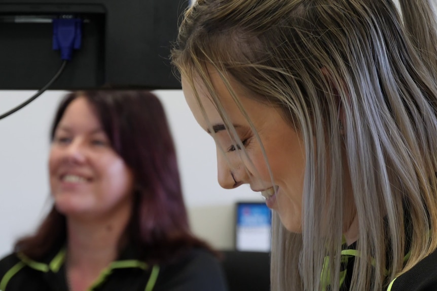 A blonde woman laughs while looking down at her computer