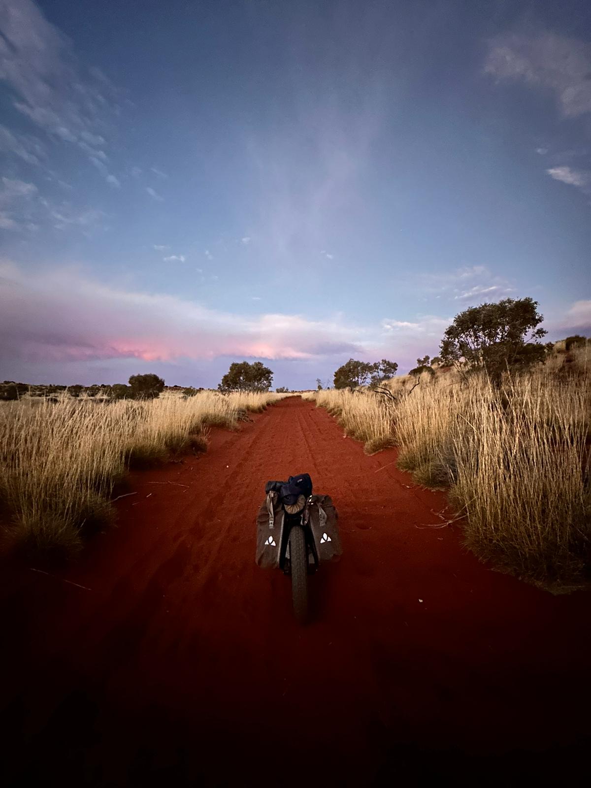 A bike in the middle of the desert, red dirt, blue sky with a tinge of pink in clouds, brown long grass on each side of path.