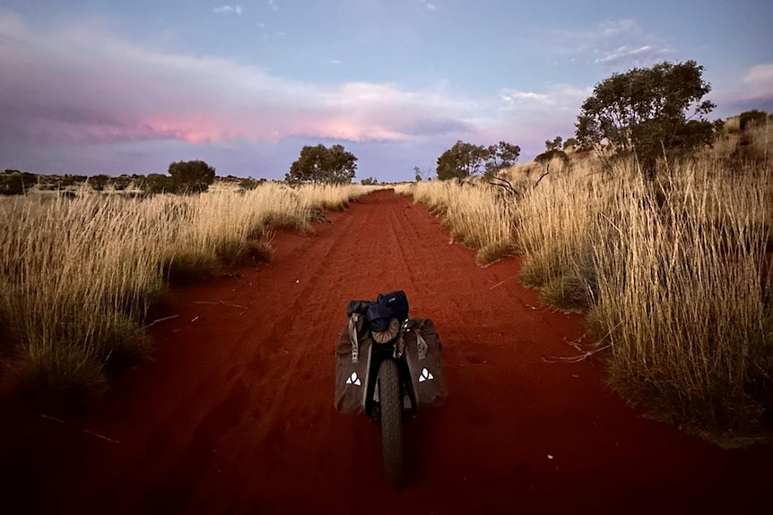 A bike in the middle of the desert, red dirt, blue sky with a tinge of pink in clouds, brown long grass on each side of path.