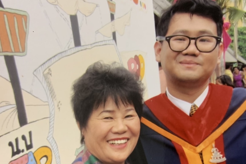 A young man in graduation robes stands next to a smiling older woman