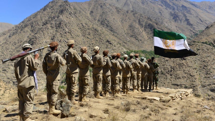 Men with weapons stand in a line, a Afghan resistance flag flies in the background