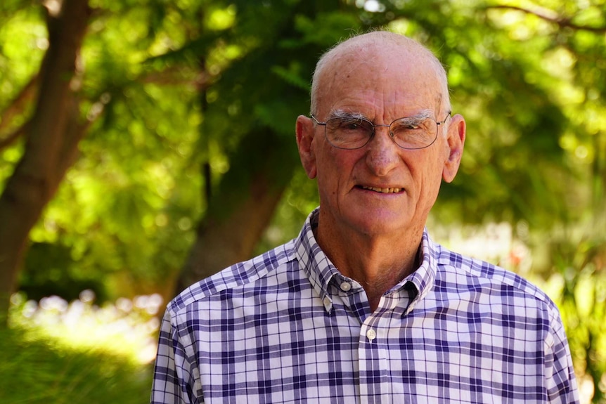 A balding man with glasses wearing a white checked shirt standing in front of some trees.