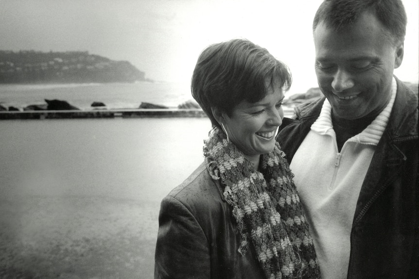 Black and white photo of couple with arms around each other and harbour in background.
