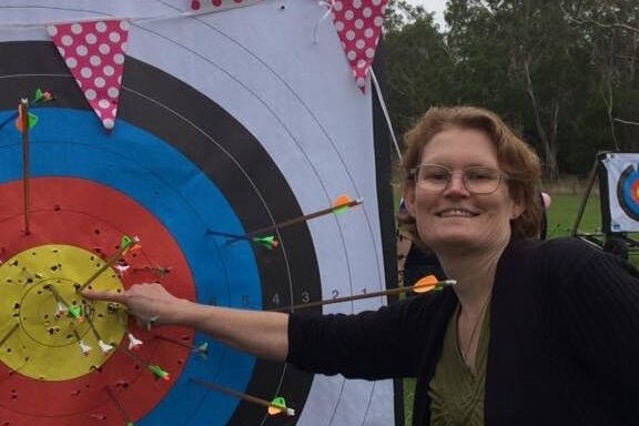 Nicole stands beside an archery board and points to an arrow embedded in the centre circle