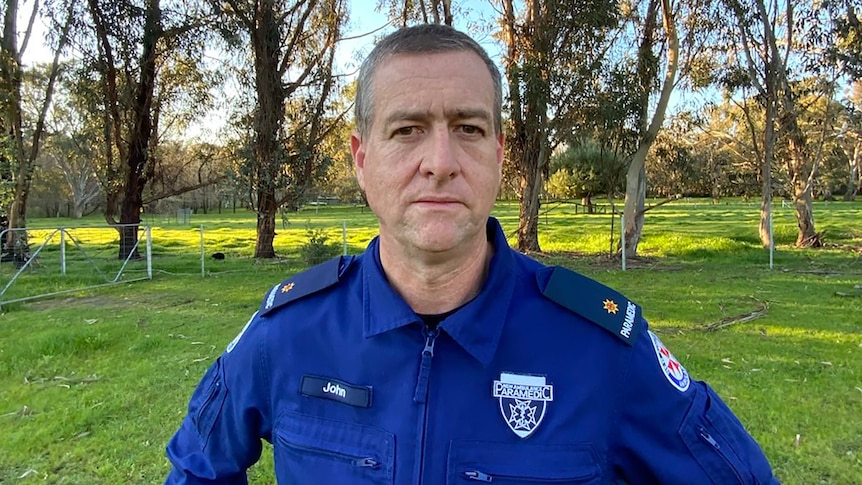 A paramedic in dark coveralls stands with his hands on his hips in a park.