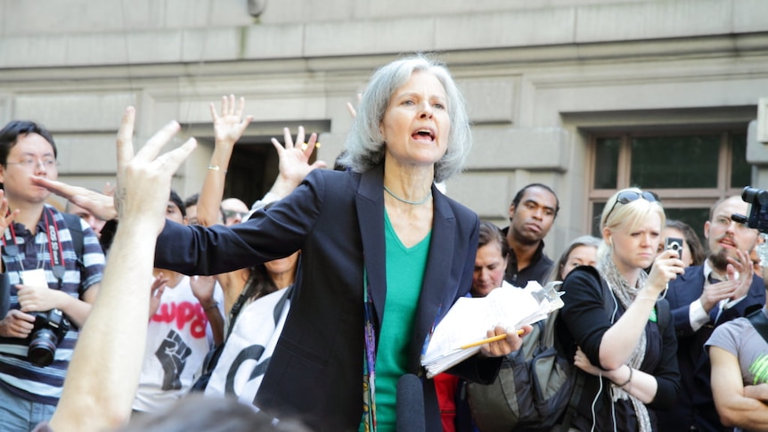 A woman with silver hair stands in a crowd, speaking to them
