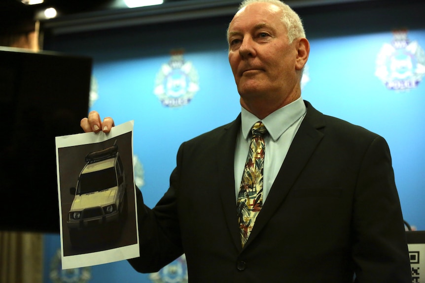 A man with white hair looks serious while holding a photo of a car.