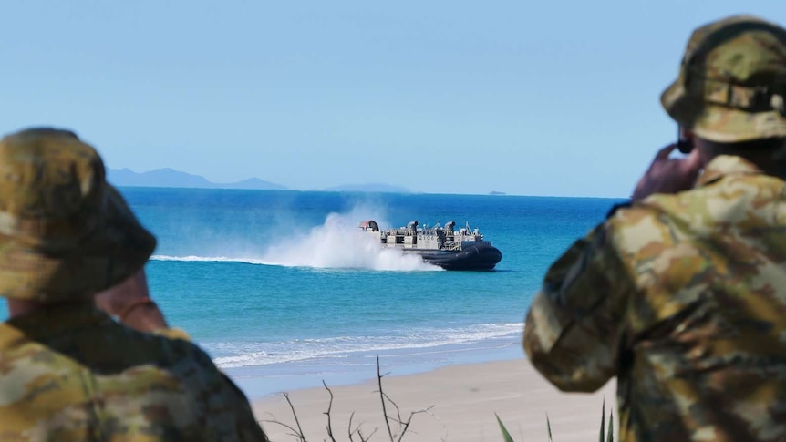 Two men in army uniforms look out to the ocean where a navy ship is approaching the shore.