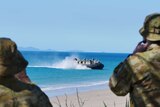 Two men in army uniforms look out to the ocean where a navy ship is approaching the shore.