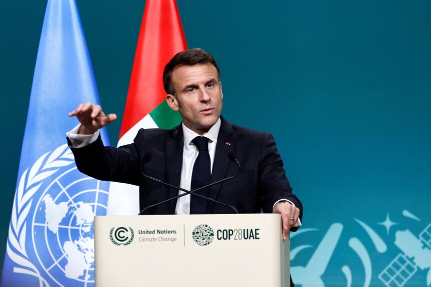 french president macron standing speaking at a podium with UN and UAE flags behind him