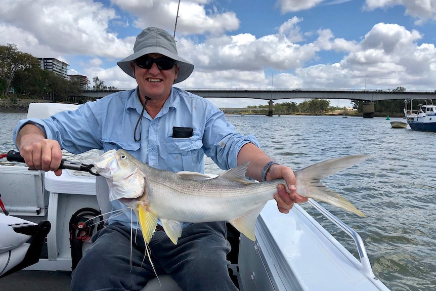 Dr Andrew Carll, wearing a hat and sunglasses, smiles as he sits on a boat in the Fitzroy River in Rockhampton.