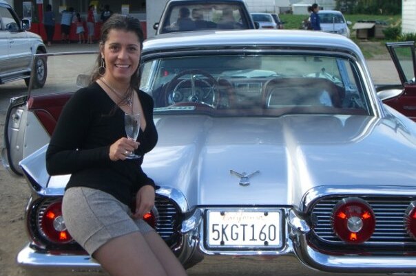 A woman with a black top holding a glass of champagne leaning on a silver classic car.