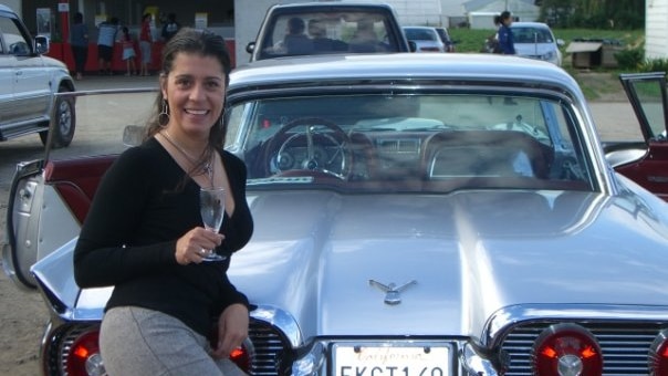 A woman with a black top holding a glass of champagne leaning on a silver classic car.