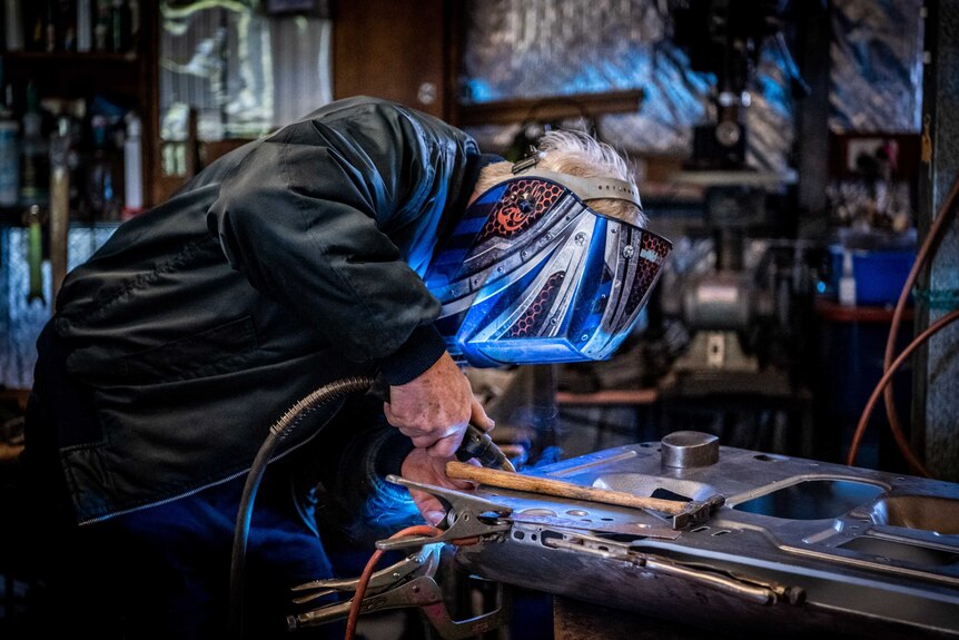 Greg Keenan wears a welding mask which reflects the blue light a blow torch as he works on a car.