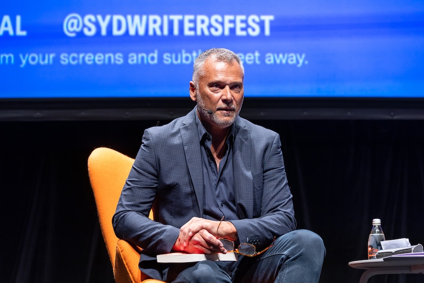 Stan Grant in a blue suit, holding a book on his knee in a yellow chair on a stage at the Sydney Writers' Festival