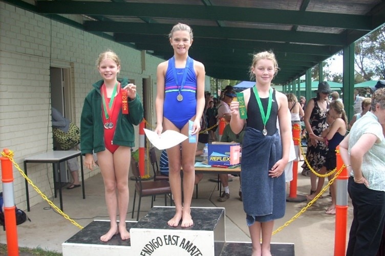 Three young girls in swimsuits stand at podium with medals around necks, one has a towel around waist, another green jacket.