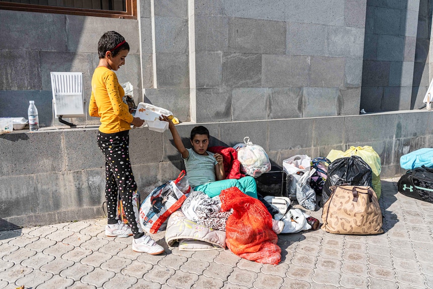 A girl hands a container to another girl who sitting on a pile of blankets and bags.