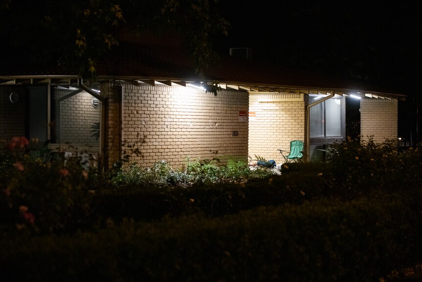 A brick building at night with some lights on, and a camping chair in the doorway.