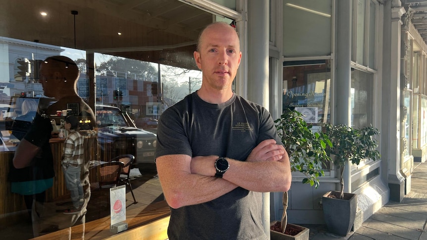 A serious bald man stands in front of a glass-fronted shop with arms crossed, wears black tee, watch, plants in shop doorway.