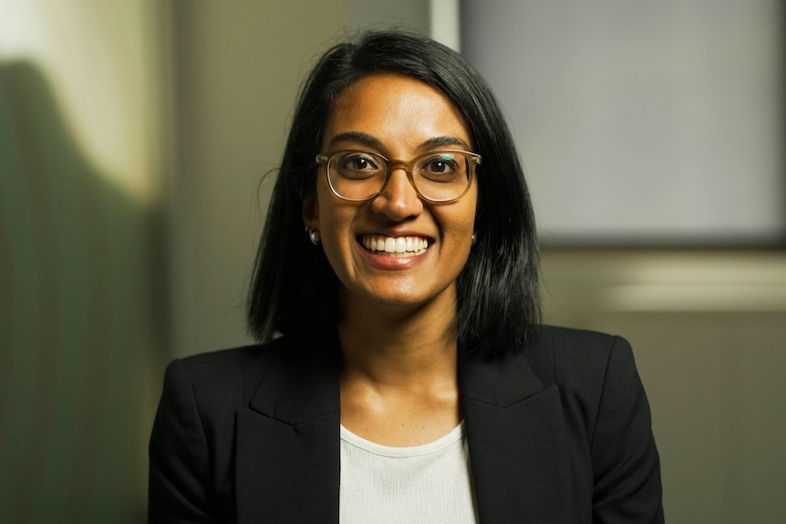 A young woman of south Asian background sitting in a chair and smiling. She's wearing glasses and has long black hair