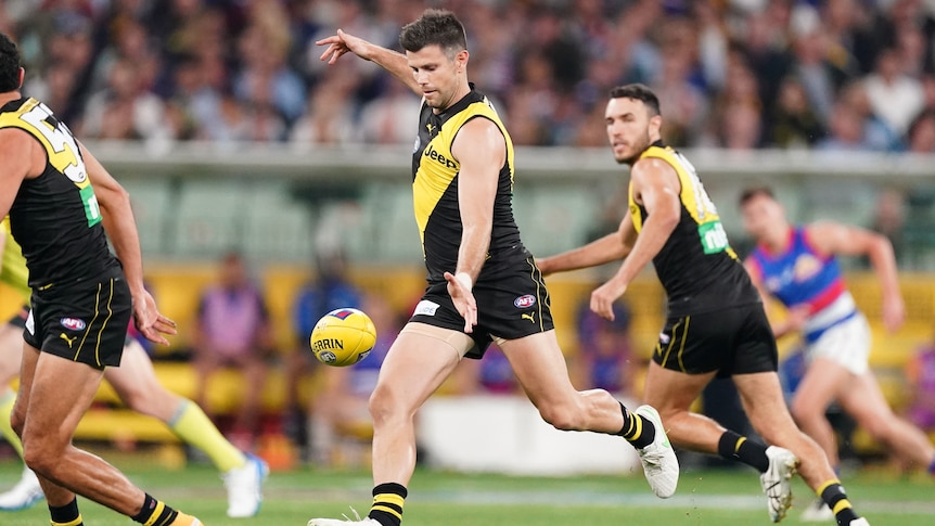 An AFL footballer drops the ball to kick it forwards as he runs down the ground with teammates surrounding him. 