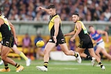 An AFL footballer drops the ball to kick it forwards as he runs down the ground with teammates surrounding him. 