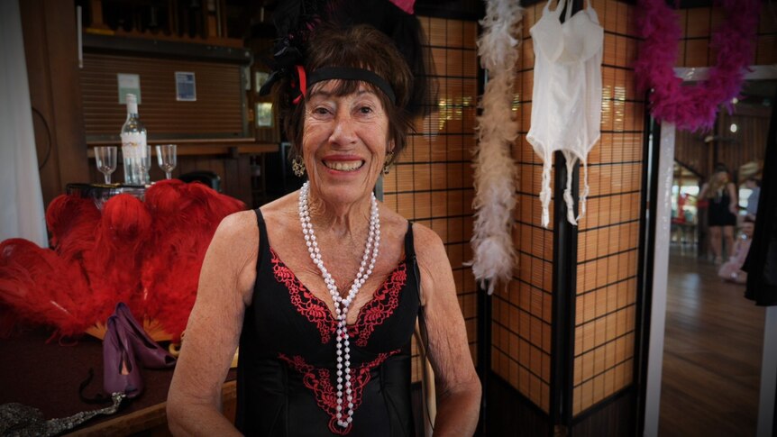 Woman in her 70s wearing a lingerie top and string of pearls, smiles, with feather boas behind her. 