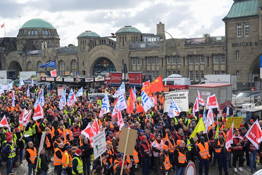 A large group of people I pictured from afar while wearing high-vis and waving flags. In the background is a large old building.
