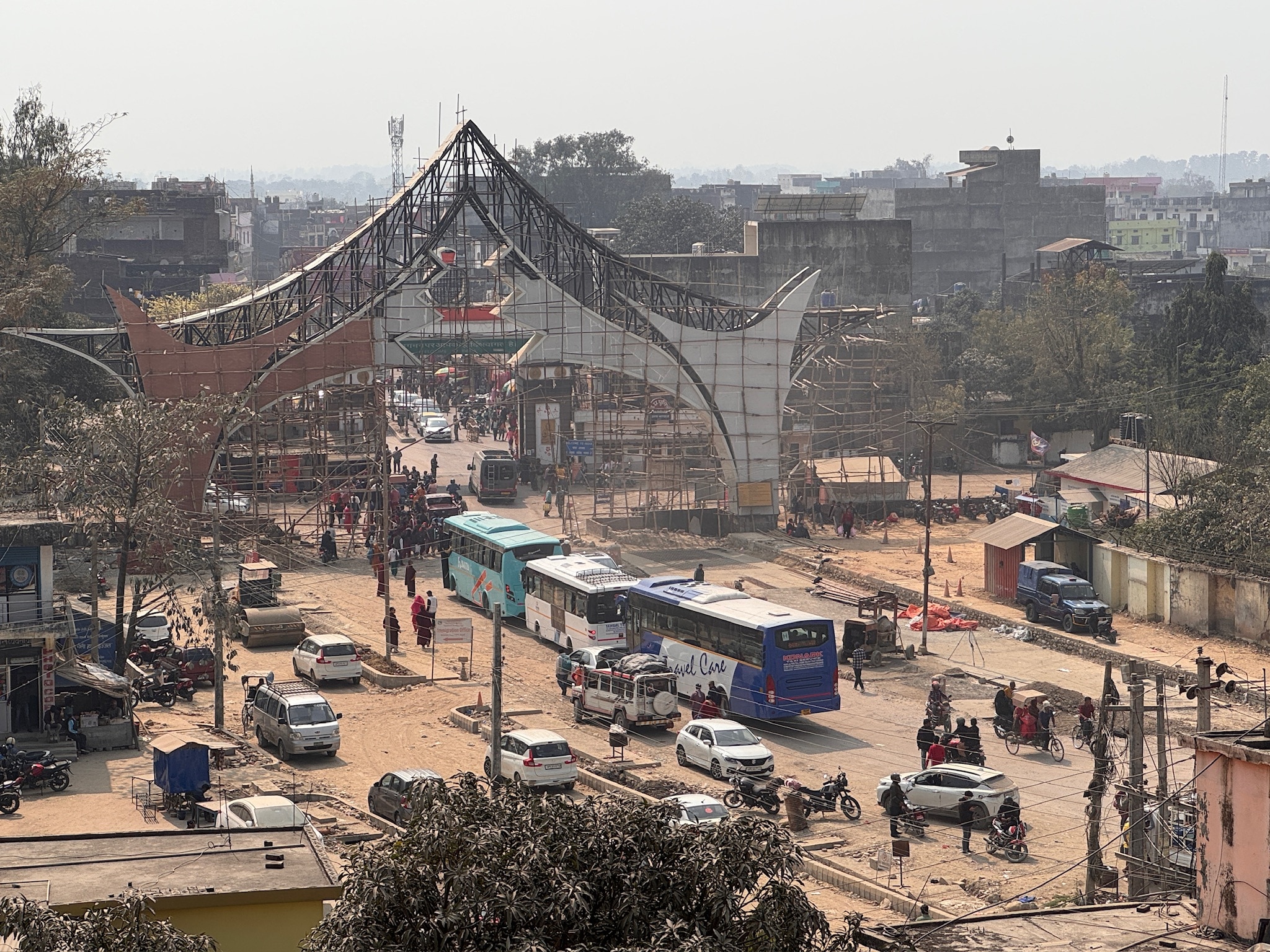 Uma grande estrutura em arco sobre uma estrada de terra com ônibus e ônibus fazendo fila com uma pequena cabana azul na beira da estrada. 