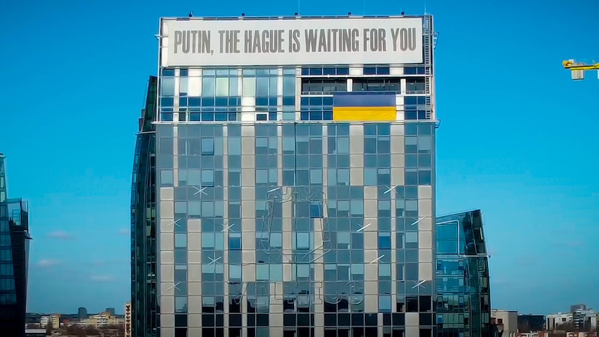 A tall office building bears a huge white banner across the top that reads 'Putin, the Hague is waiting for you'