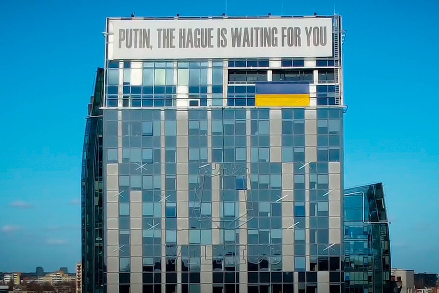 A tall office building bears a huge white banner across the top that reads 'Putin, the Hague is waiting for you'