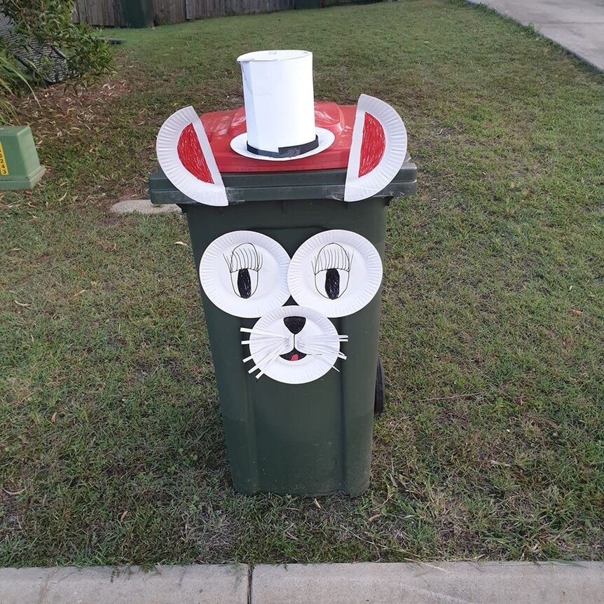 A kerbside rubbish bin with bunny ears, a nose, eyes and a top hat all made out of white cardboard