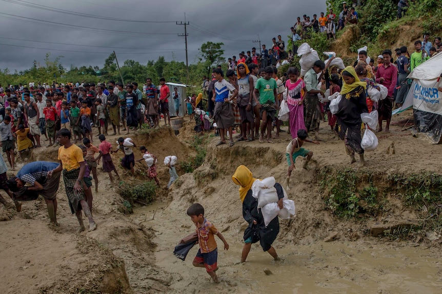 A crowd of people walk carrying aid through a muddy refugee camp.