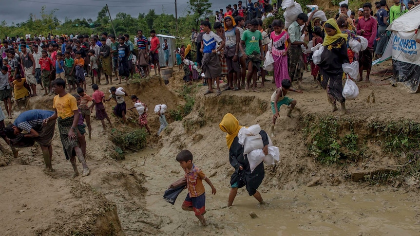 A crowd of people walk carrying aid through a muddy refugee camp.