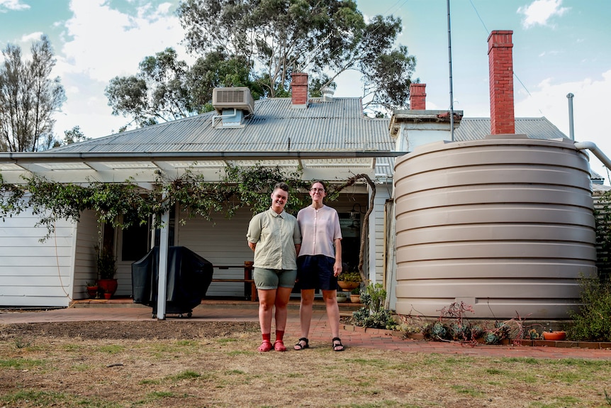 Two woman standing outside a home with a large water take and trees visible in background 